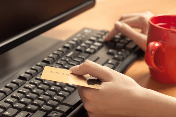 Computador com teclado e xícara de café vermelha, com um cartão de crédito em uma das mãos e a outra simulando o ato de digitação | Comprar gift cards de forma segura.